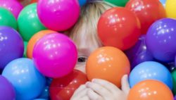 Foto: Ein Kindergesicht ist zwischen vielen bunten Bällen in einem Bällebad erkennbar