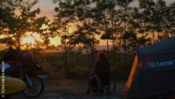 Foto: Rollstuhlfahrerin steht zwischen einem Zelt und einem Motorrad und beobachtet den Sonnenuntergang; Copyright: TOSHI MEIER-BROOK